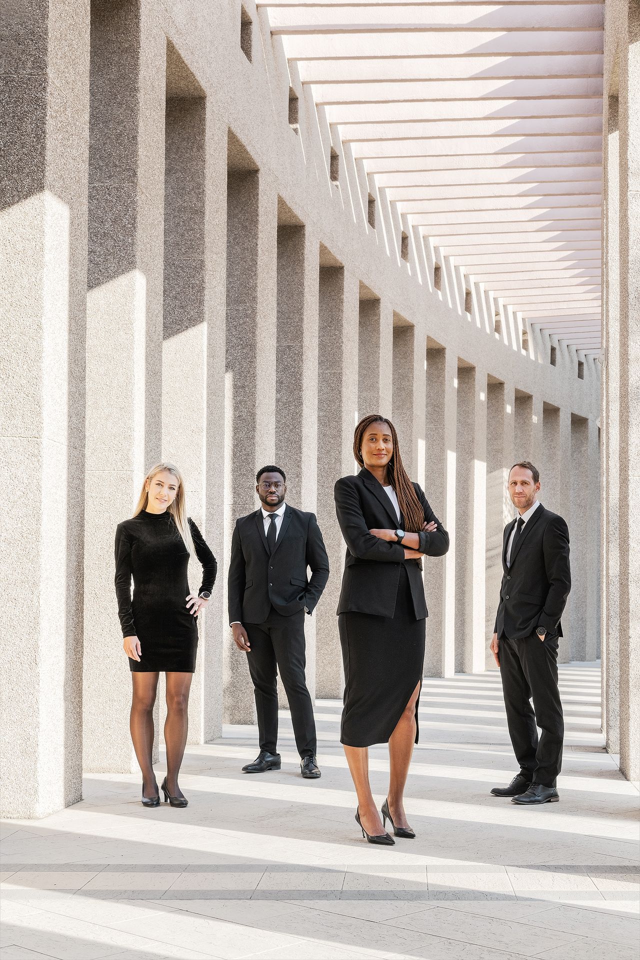 Four diverse business professionals in black attire stand confidently in a modern columned corridor.