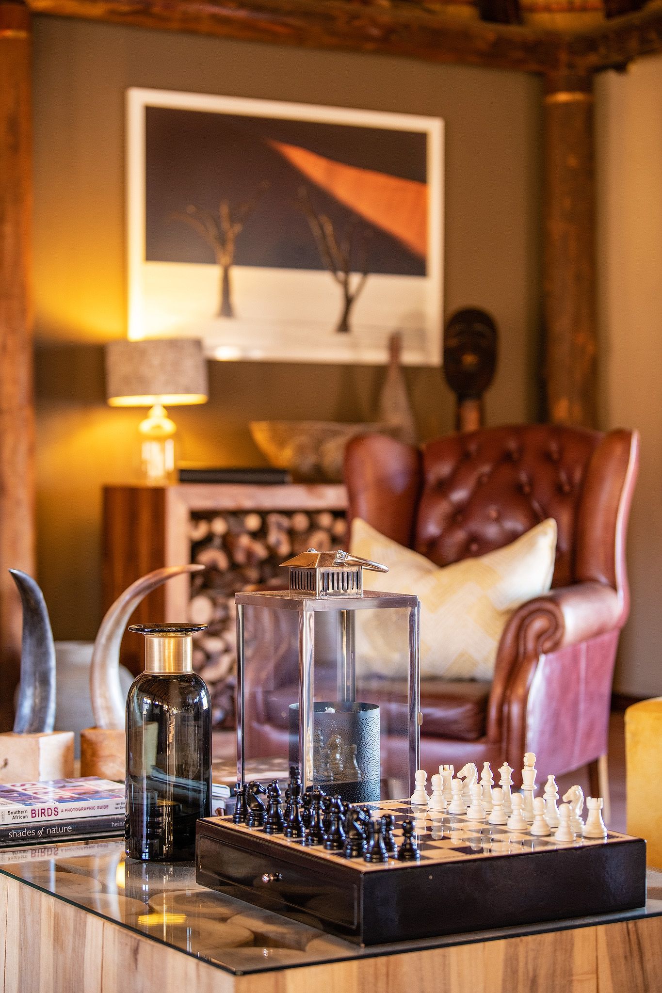 Cozy safari lodge lounge with chess board, glass lanterns, leather armchair, and African-inspired artwork on the wall.