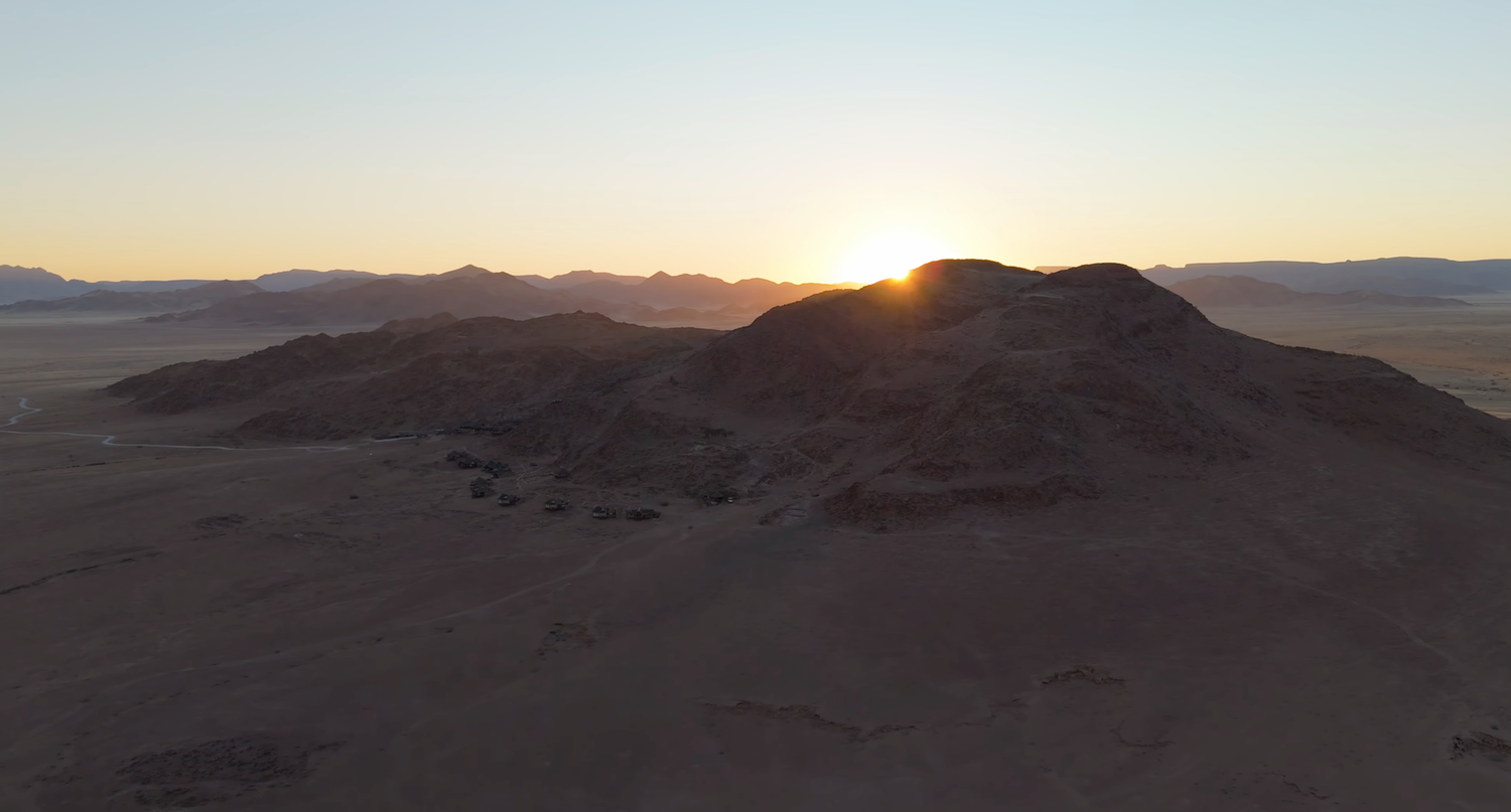 Aerial view of a rocky desert hill silhouetted against a golden sunset with mountain ranges in the background.