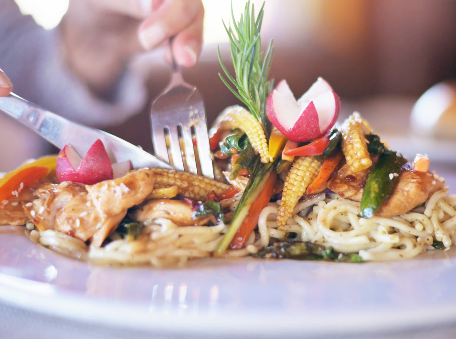 Gourmet noodle dish with grilled chicken, baby corn, fresh vegetables, and herbs served on a white plate.
