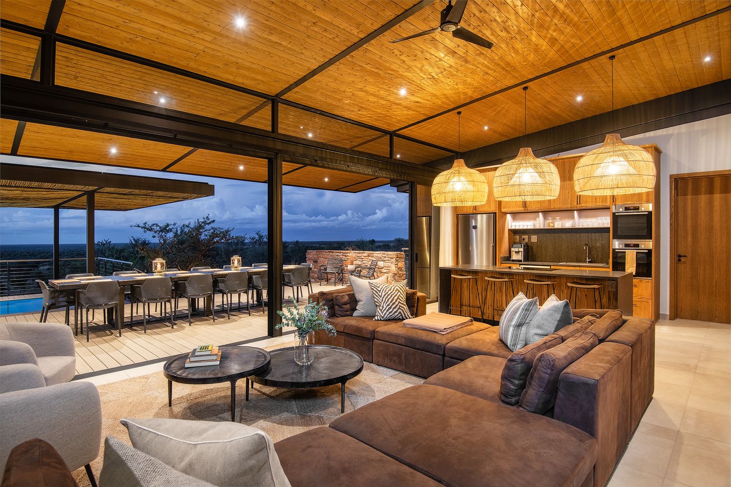 Open-plan luxury safari villa living area with thatched roof, pool, and panoramic bush views at dusk.