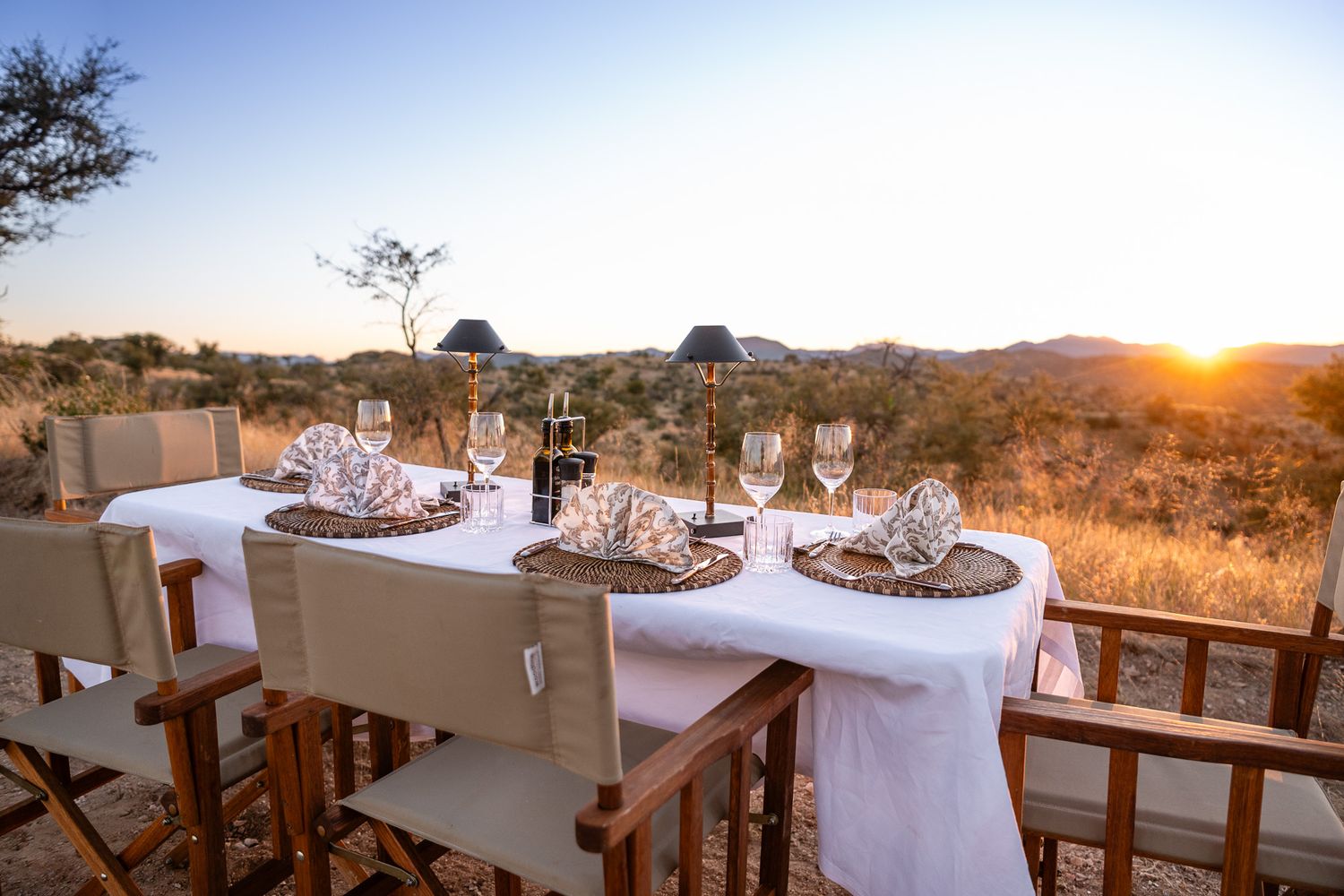 Elegant outdoor safari dining table set for sunset dinner with wine glasses and woven placemats overlooking African savanna landscape.