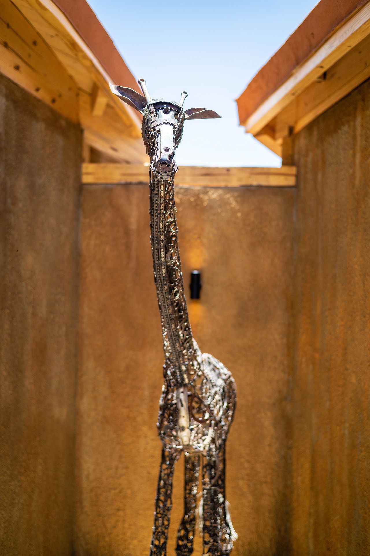 A tall metallic giraffe sculpture stands between two rustic wooden structures against a clear blue sky.