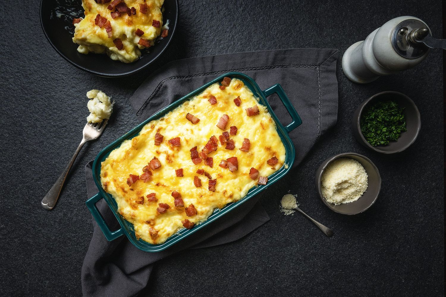 Golden baked cauliflower mac and cheese casserole with crispy bacon in a teal dish, served on a dark slate background.