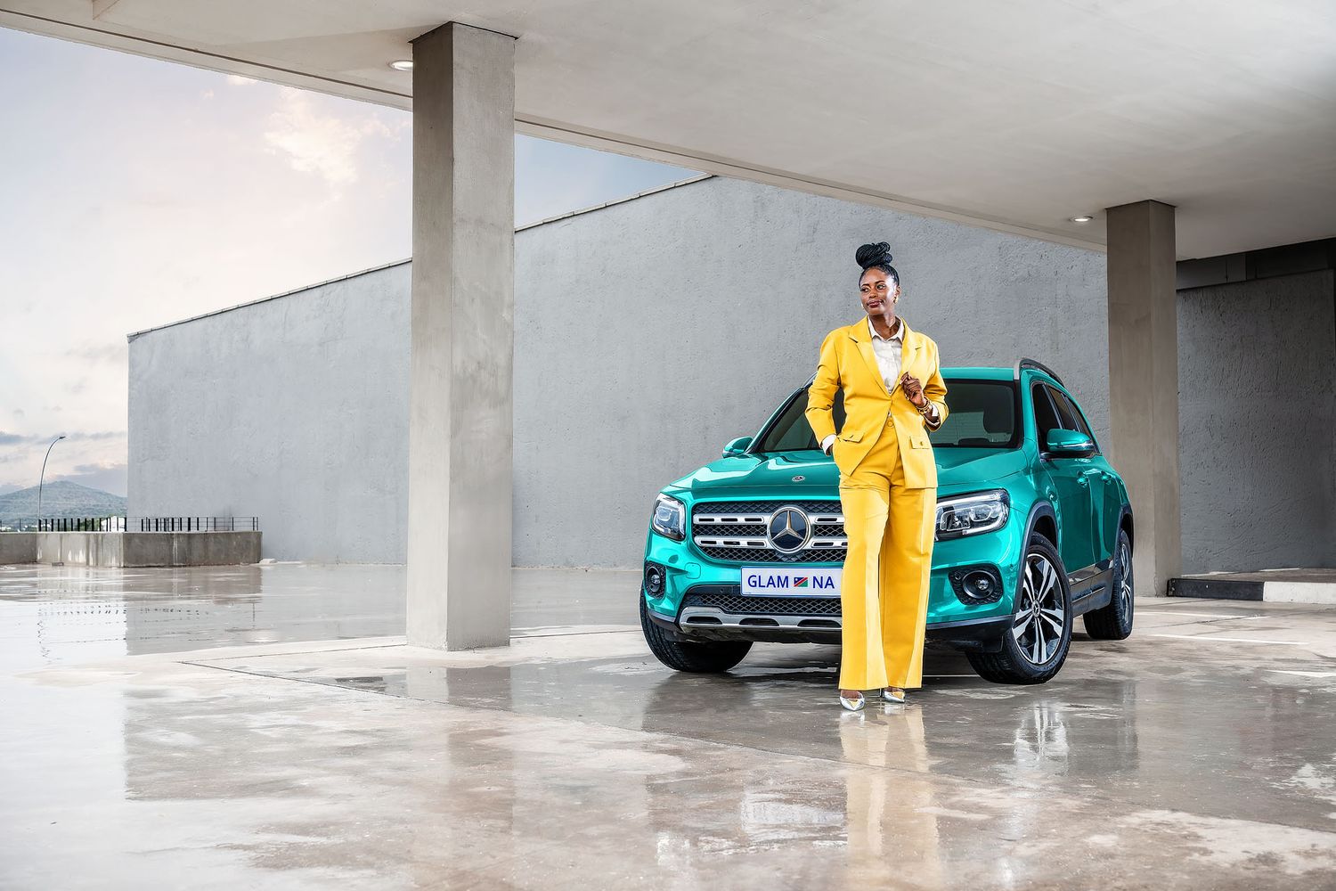 Woman in yellow pantsuit posing beside a teal Mercedes-Benz pickup truck in a modern parking structure. FNB
