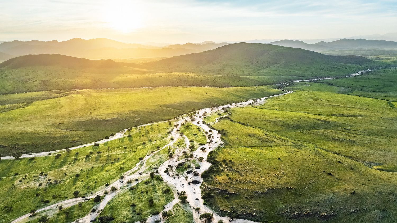 Aerial view of a winding river through lush green valley with mountains and golden sunset in the background.