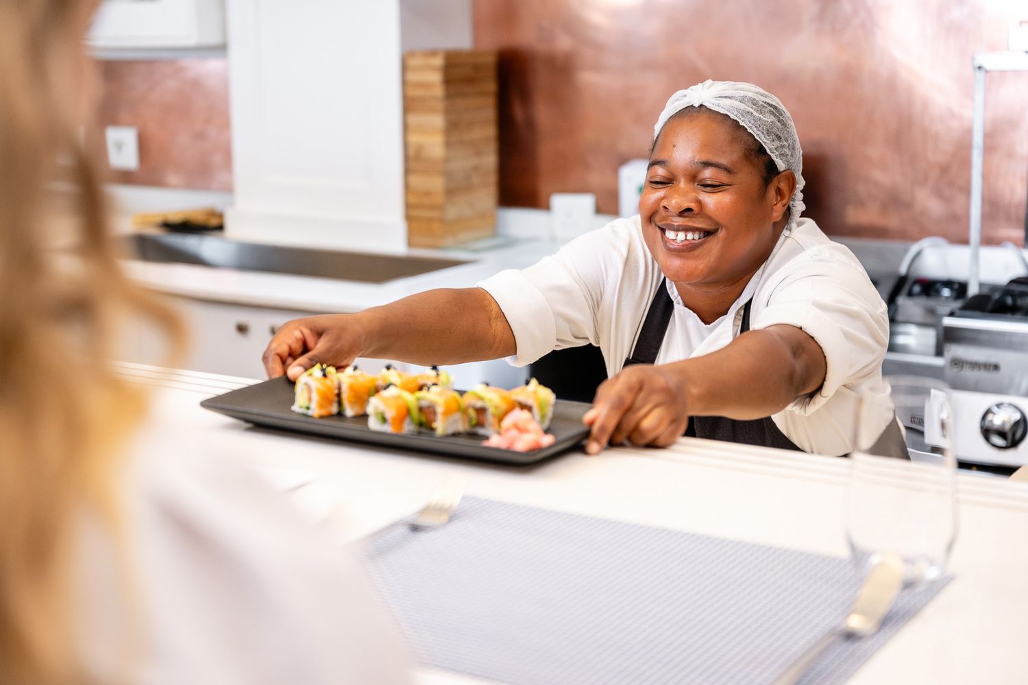 Smiling female chef in hairnet presents a sushi platter on a black tray in a professional kitchen.