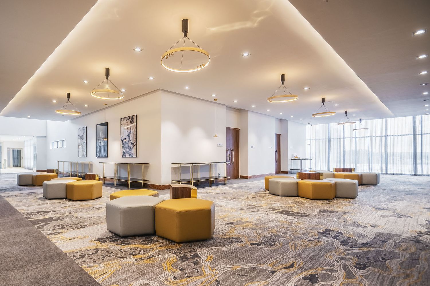 Modern hotel lobby with gold and gray modular seating, patterned carpet, circular pendant light, and floor-to-ceiling windows.