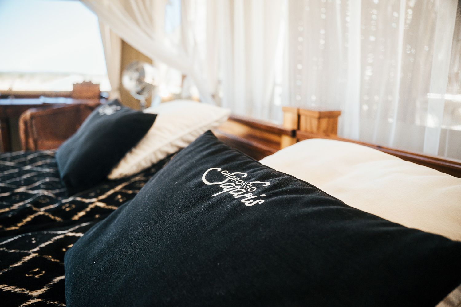 Cozy bedroom with black embroidered pillow on bed, sheer white curtains, and ocean view in background.