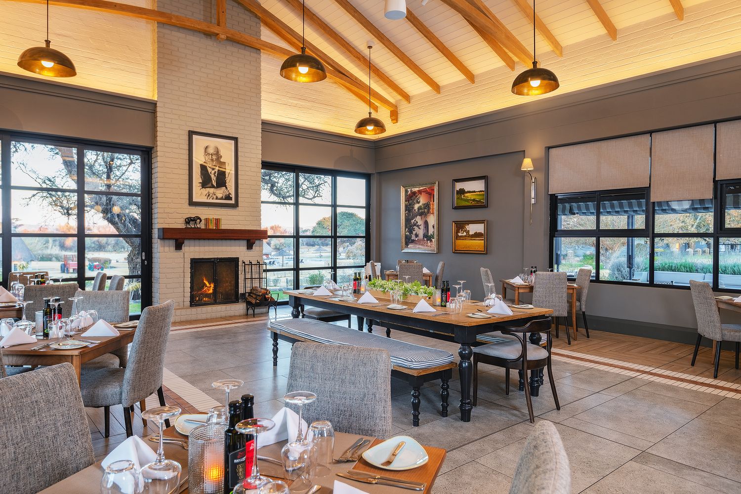 Elegant restaurant interior with fireplace, wooden ceiling, large windows, and neatly set dining tables in a warm ambiance.