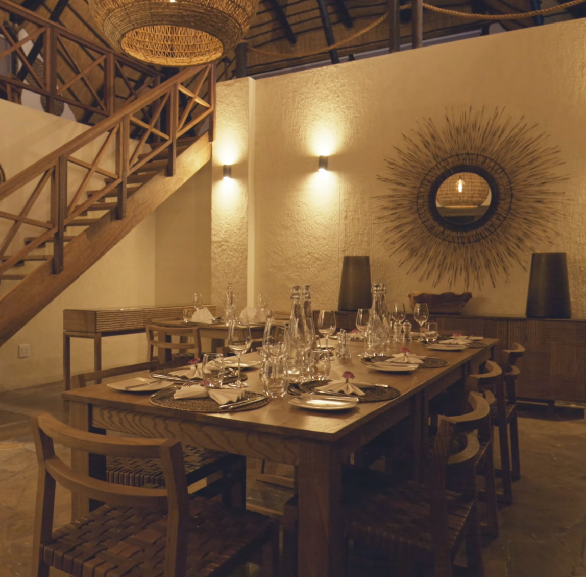 Rustic dining room with wooden table set for dinner, starburst mirror, warm wall sconces, and exposed wooden staircase.