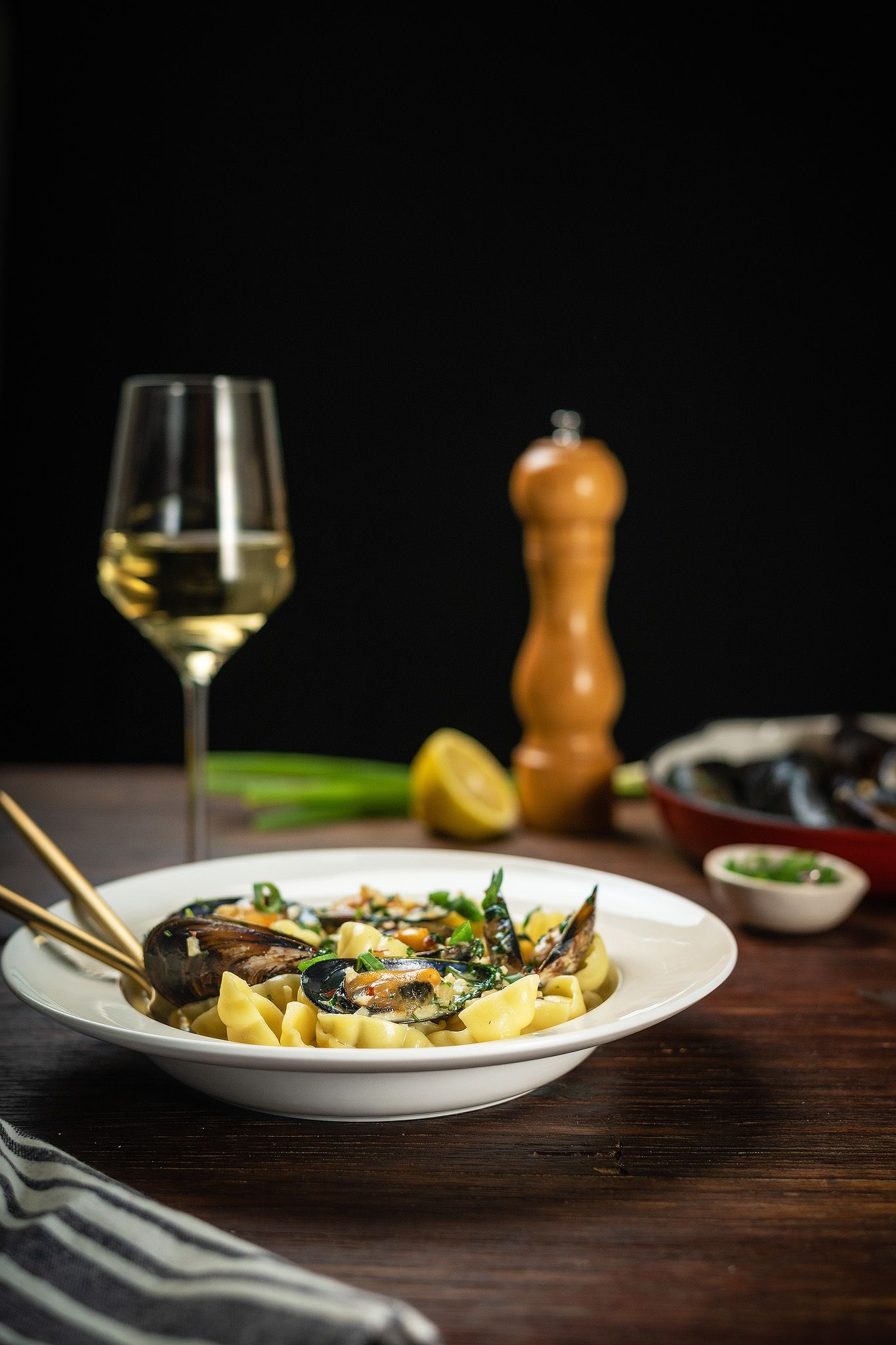 Elegant pasta dish with mussels and greens in white bowl, served with white wine and pepper grinder on wooden table.