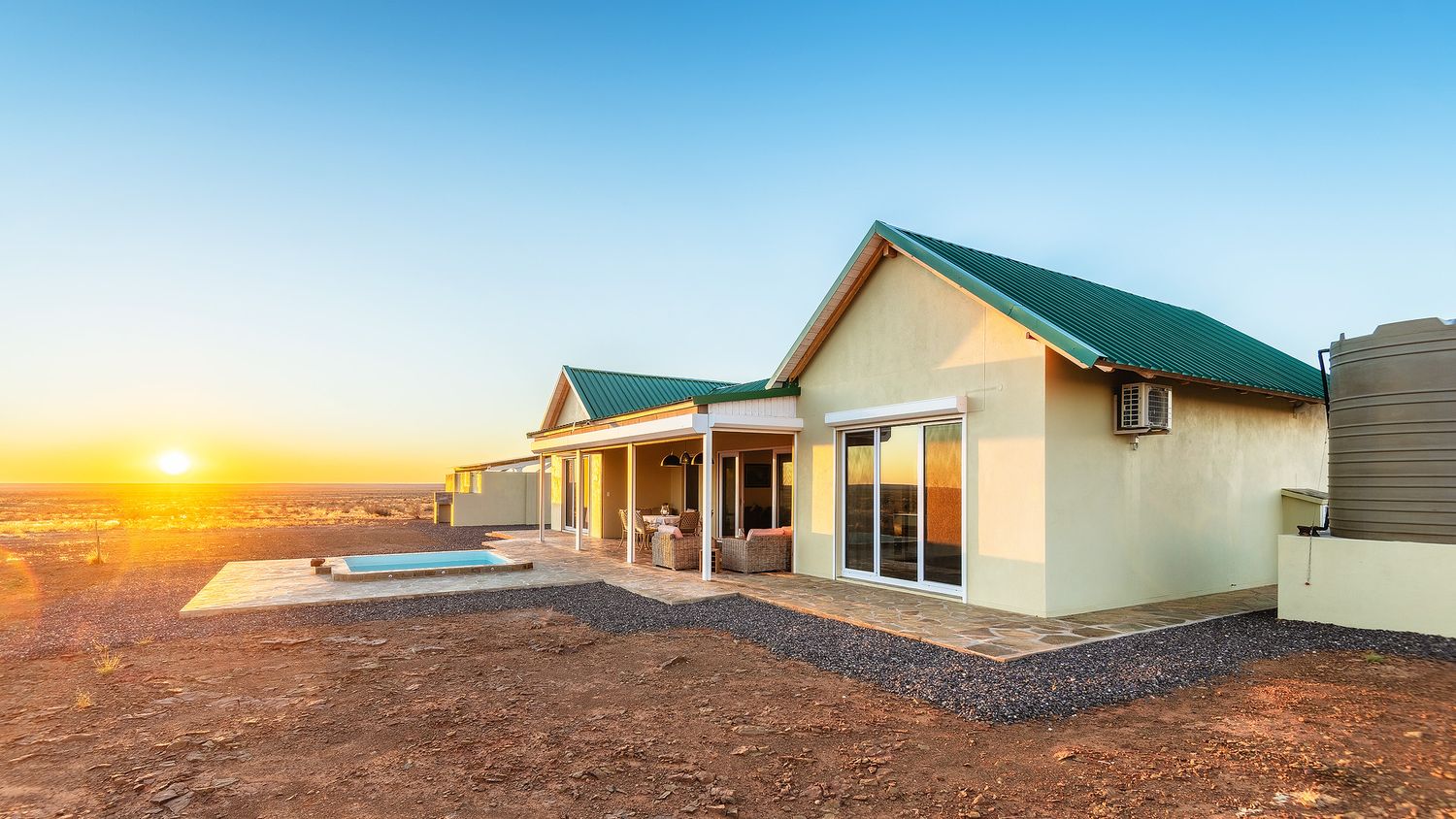 Modern farmhouse with green roof at sunset, overlooking vast arid landscape under a golden sky.