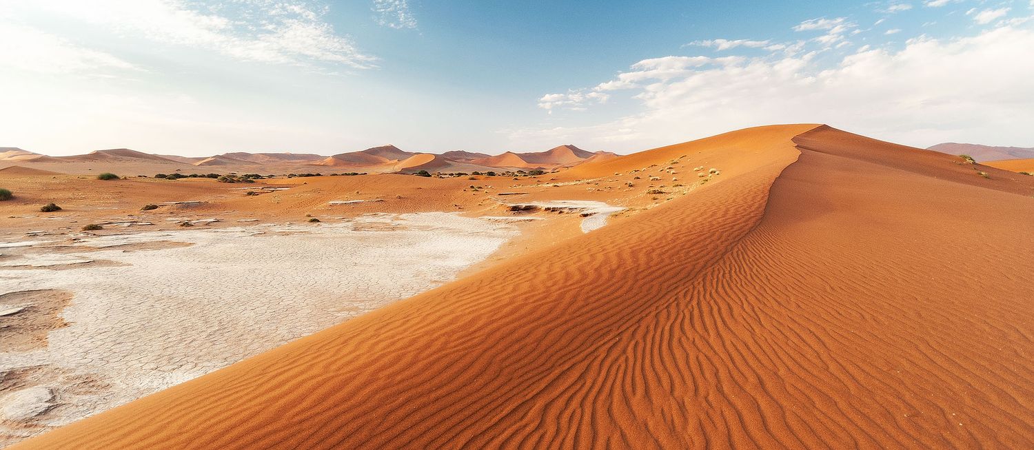 Sweeping red sand dunes of the Namib Desert stretch under a blue sky, with rippled textures and distant mountains.