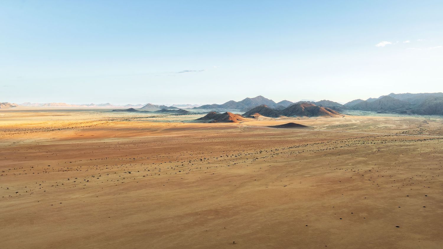 Vast desert landscape with golden sand dunes, sparse vegetation, and distant mountains under a clear blue sky.