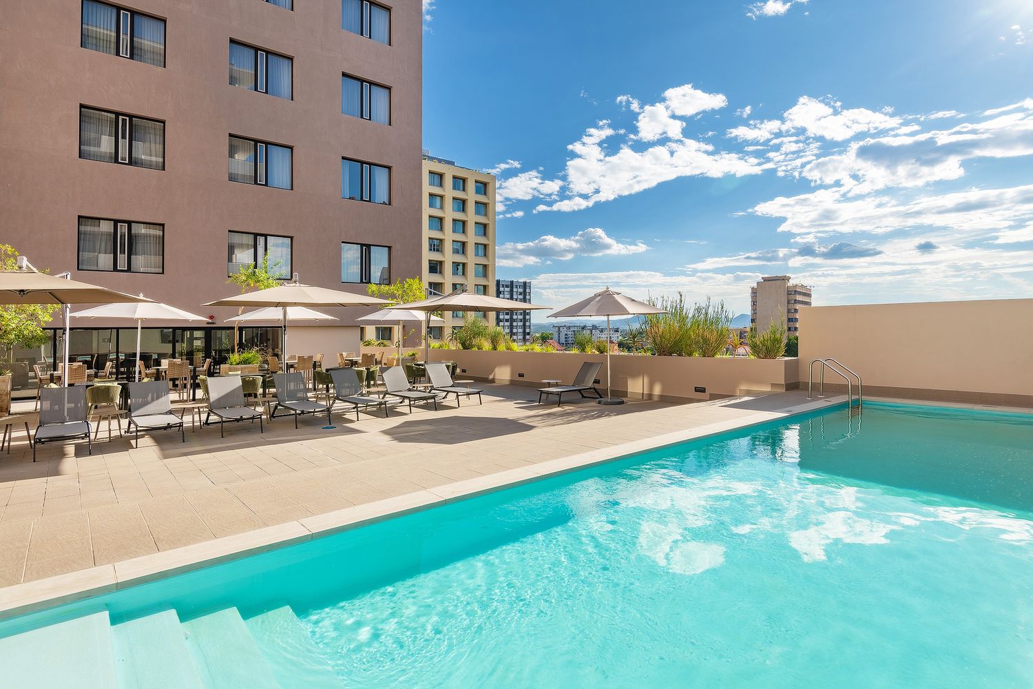 Hotel outdoor pool with lounge chairs and umbrellas, surrounded by multi-story buildings under a bright blue sky.
