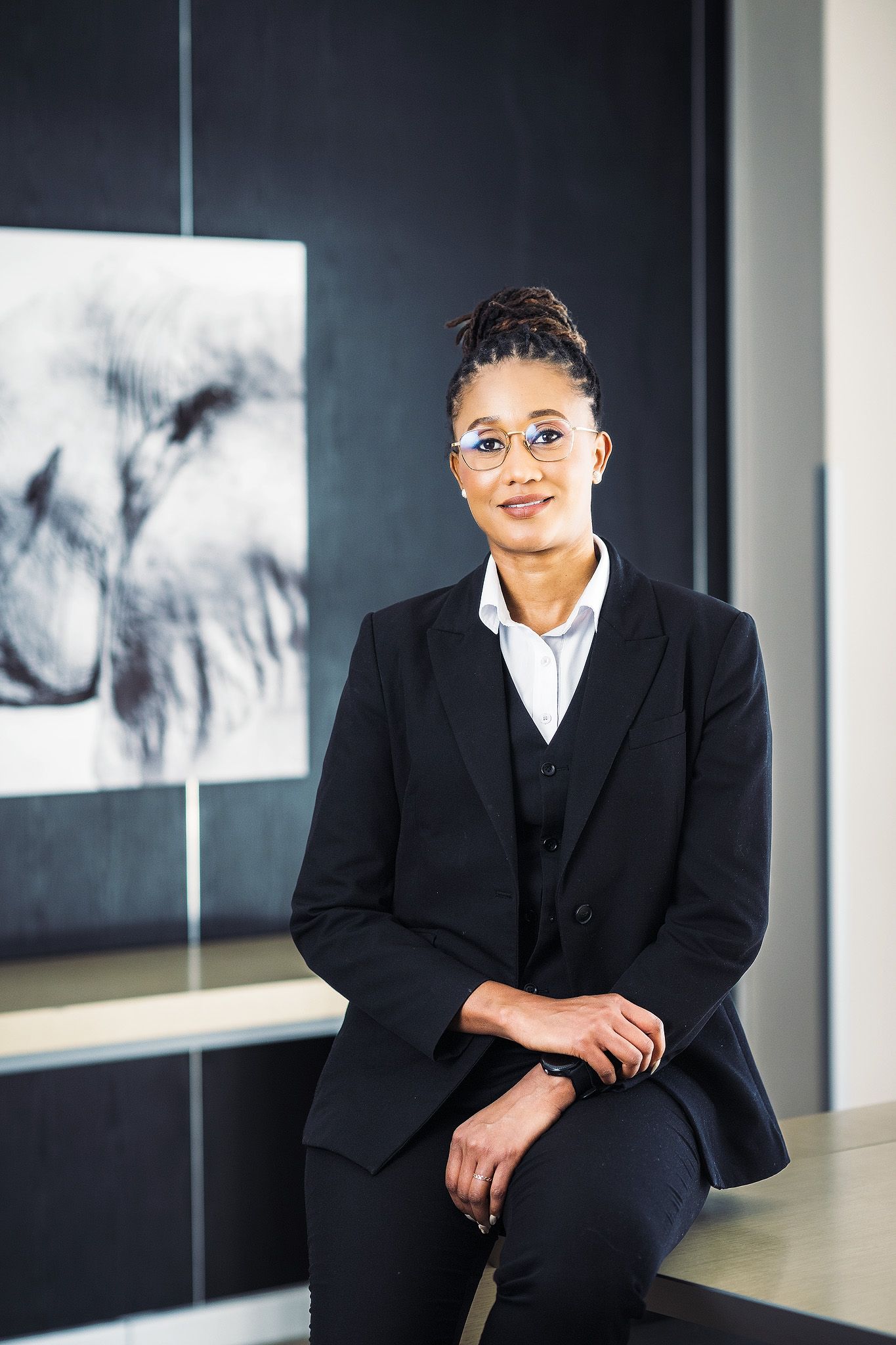 Professional woman in black blazer and glasses smiling confidently in a modern office setting.