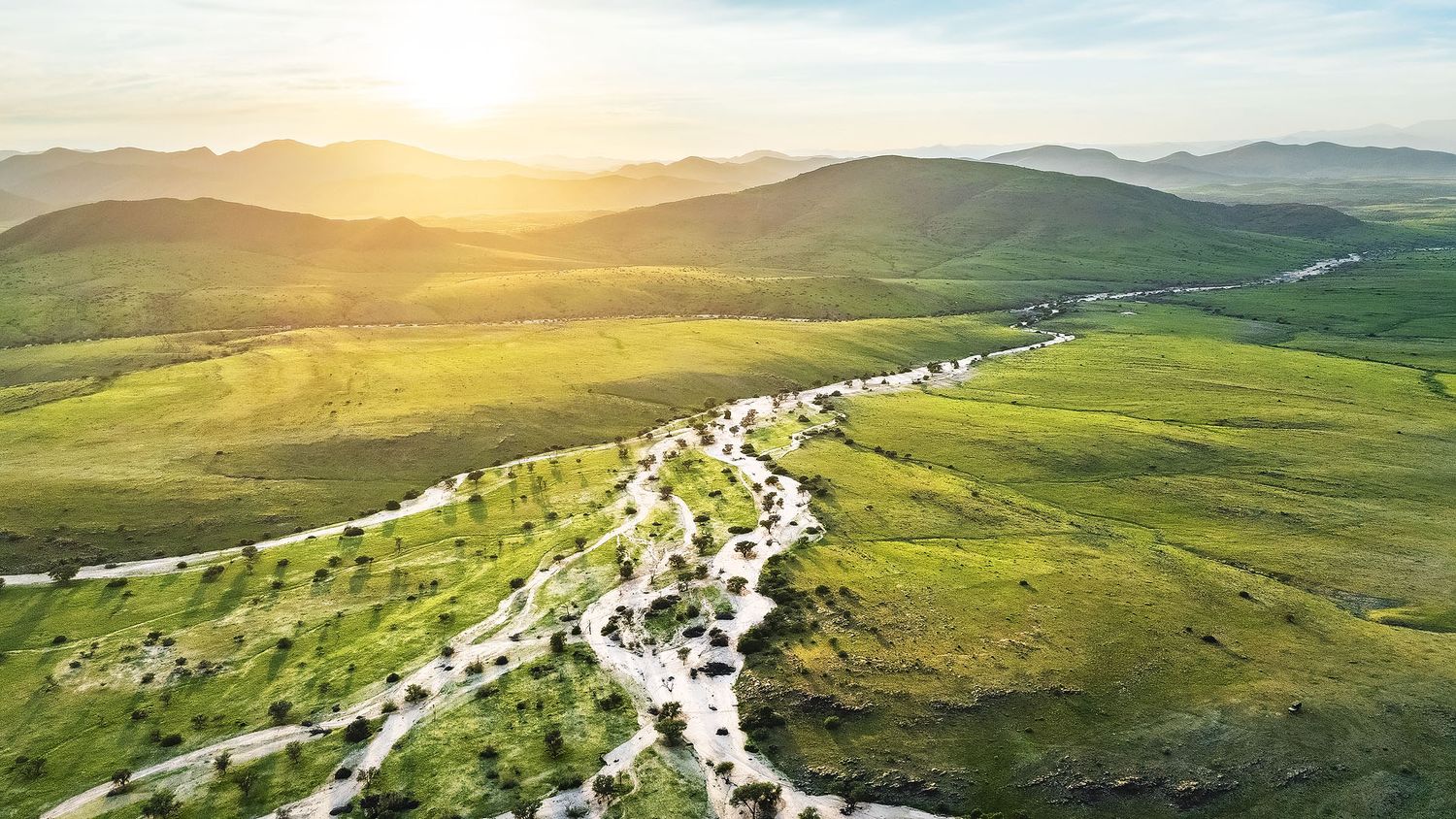 Aerial view of a winding river through lush green valley with rolling mountains and golden sunset in the background.