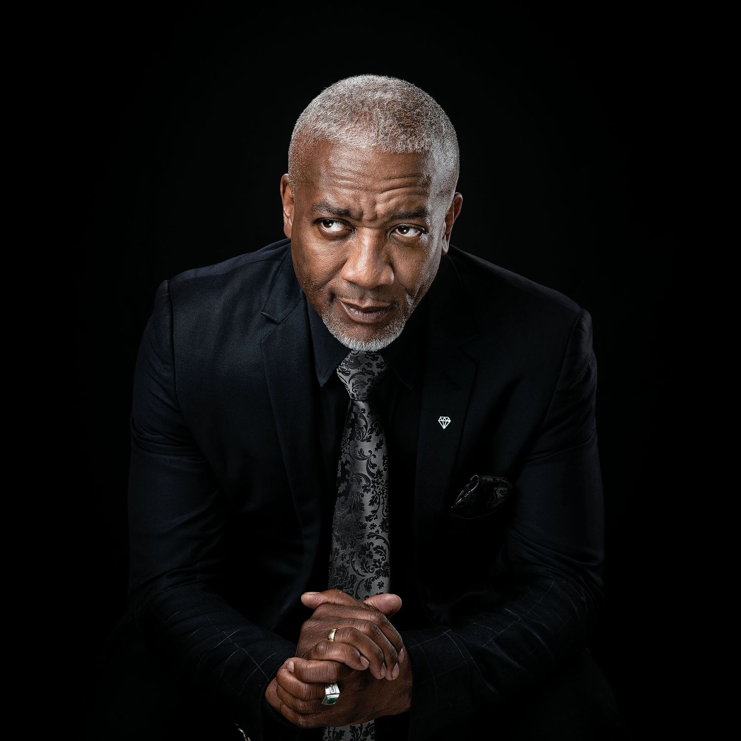 Professional portrait of distinguished older Black man in black suit and tie against dark background.