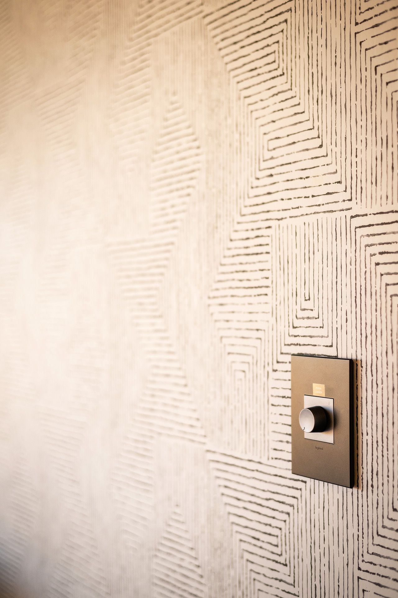 A bronze dimmer switch mounted on a textured beige wall with geometric square pattern wallcovering.