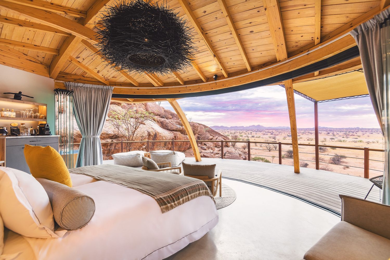 Luxury safari lodge bedroom with open walls, king bed, wooden ceiling, and breathtaking desert landscape views at sunset.