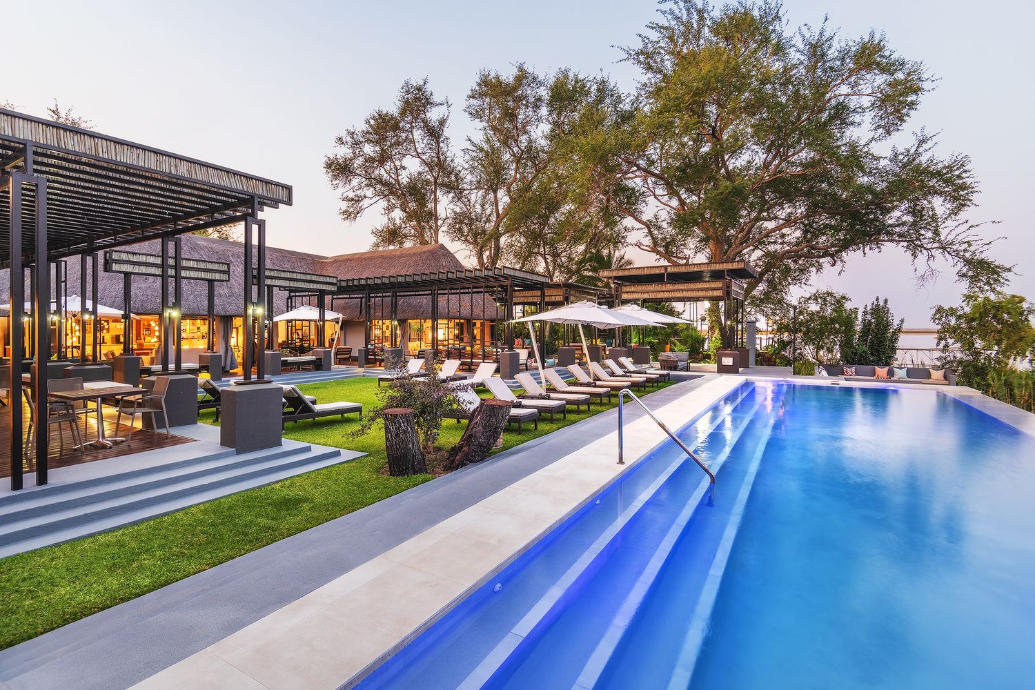 Luxury resort infinity pool with lounge chairs, lush greenery, and an elegant restaurant terrace at dusk.