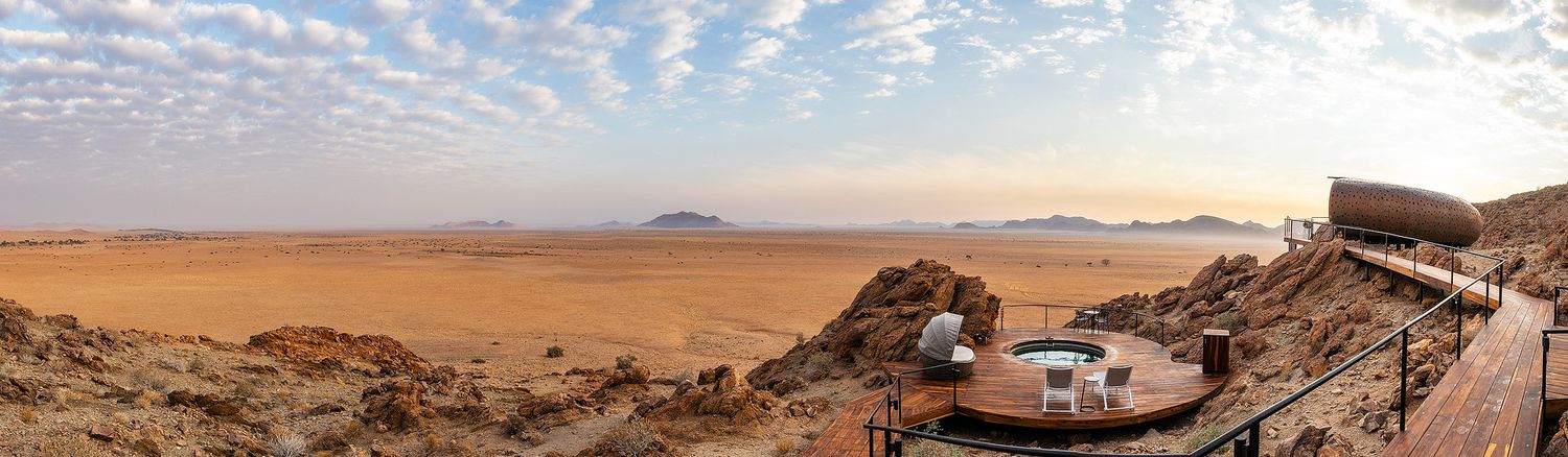 Panoramic view of vast Namibian desert landscape with rocky terrain, scattered structures, and mountains under a cloudy sky.