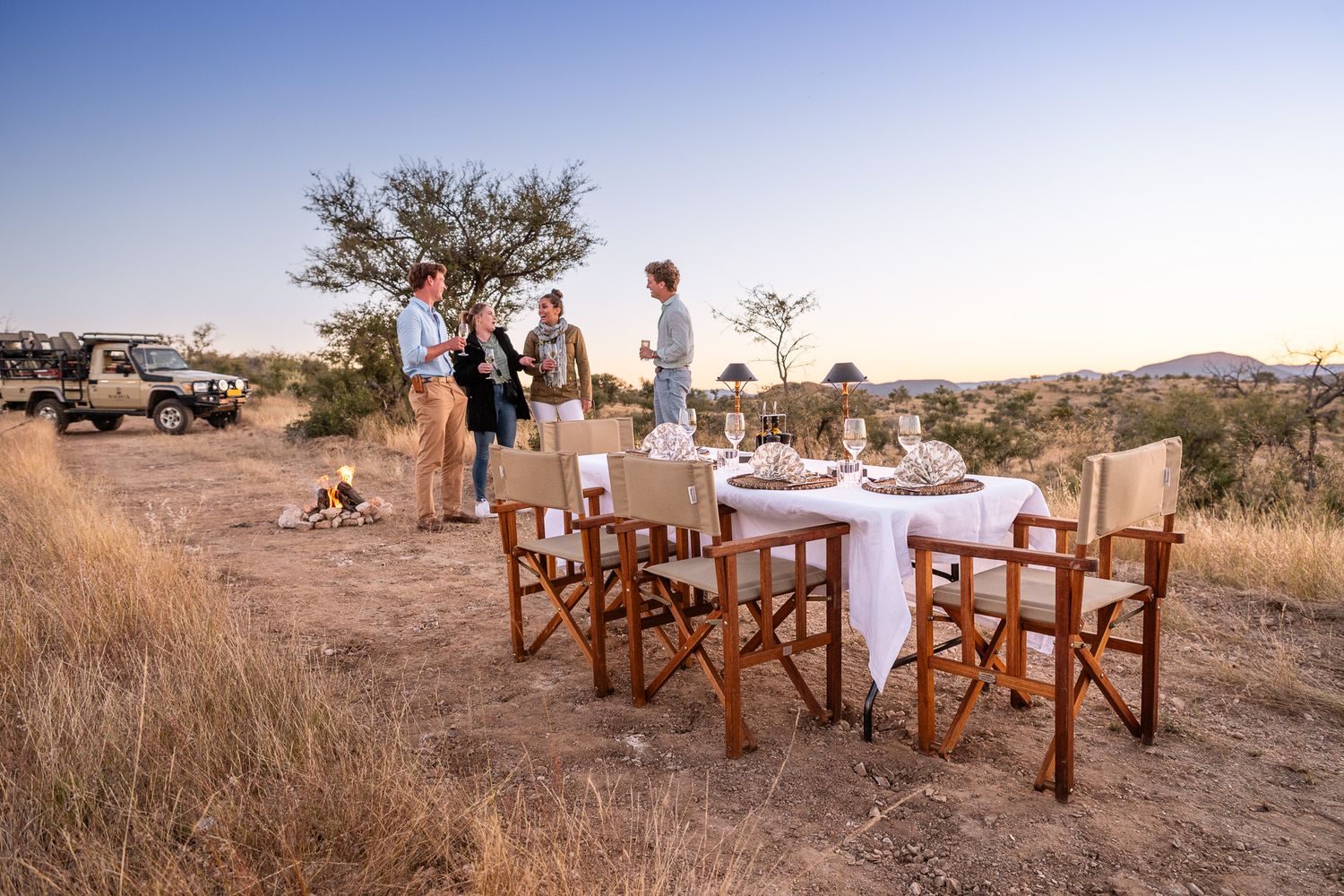 Guests enjoy a bush dinner at sunset in the African savanna with safari vehicle and acacia trees in background.