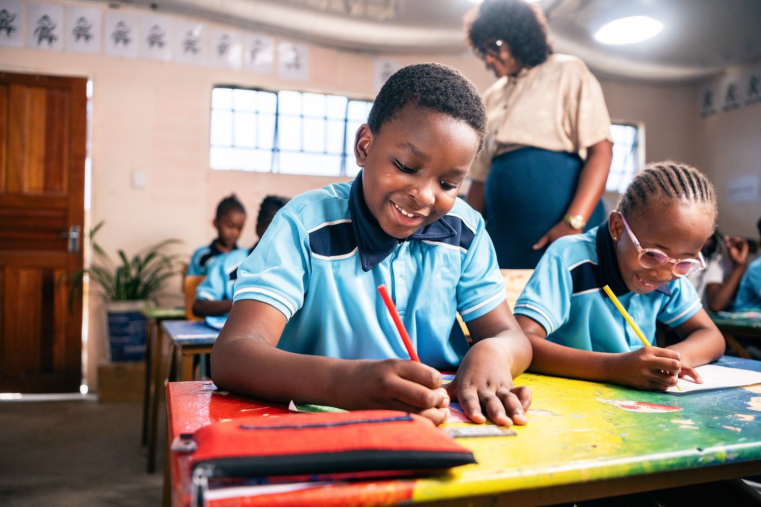 Happy African schoolchildren in blue uniforms drawing with colored pencils in a bright classroom.
