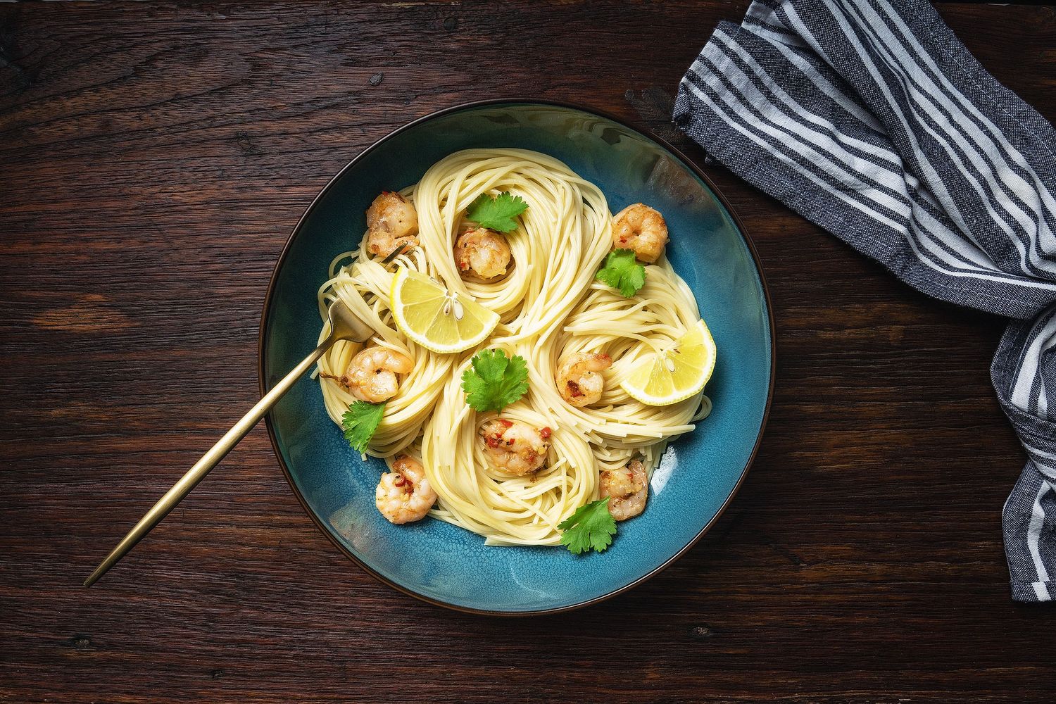 Shrimp scampi pasta with lemon slices and parsley in a blue bowl on a dark wooden table.