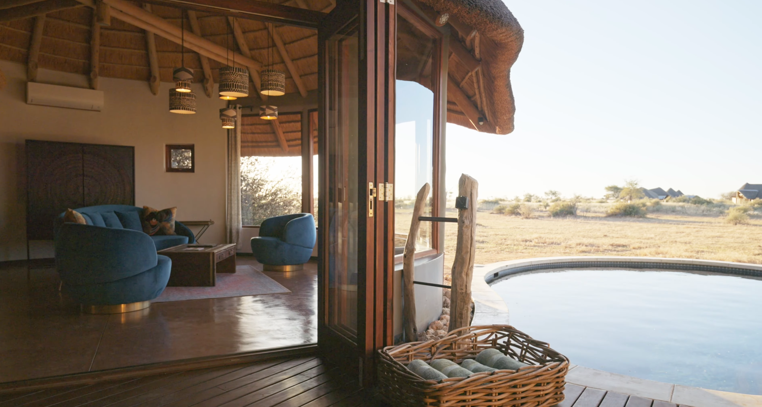 Luxury safari lodge with plunge pool, open-plan living area, blue chairs, and sweeping desert views at golden hour.