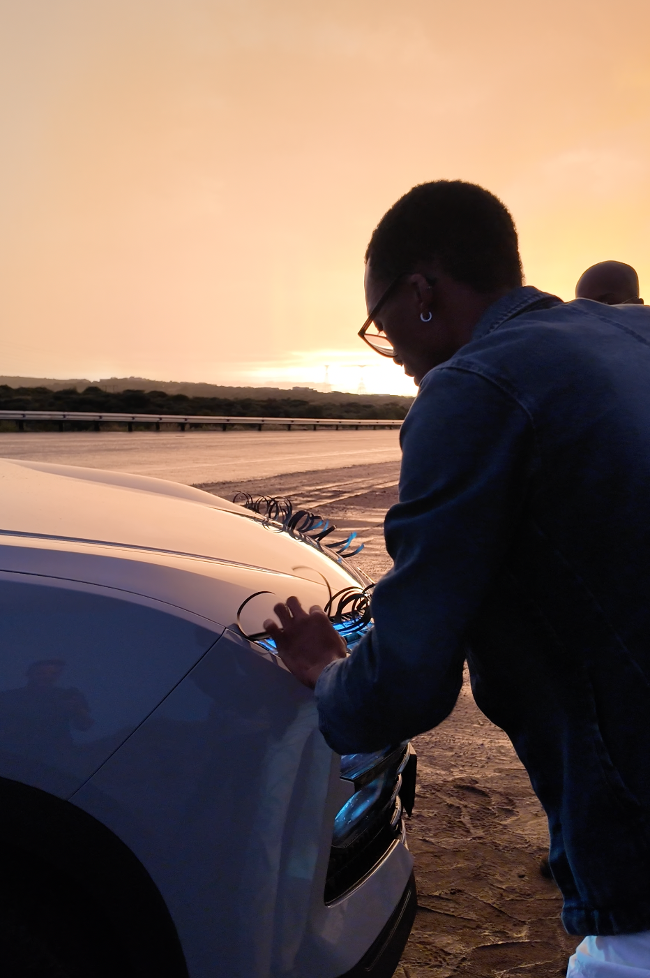 A young man leans over a white car hood at sunset, wearing a blue jacket, with a golden sky and road in the background.