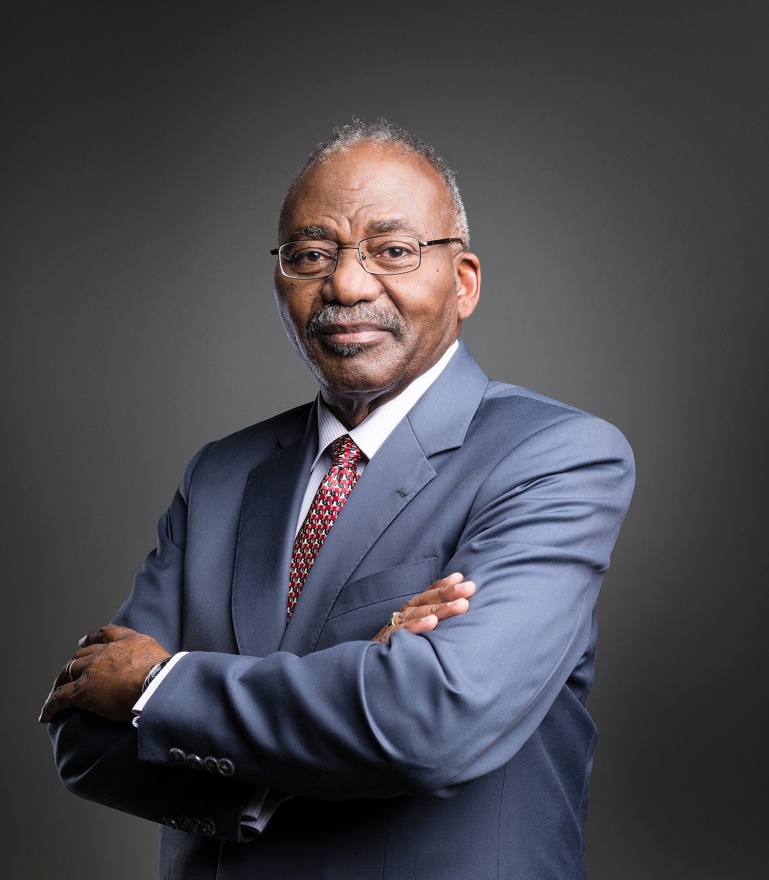 Professional businessman in blue suit with crossed arms posing confidently against gray background.