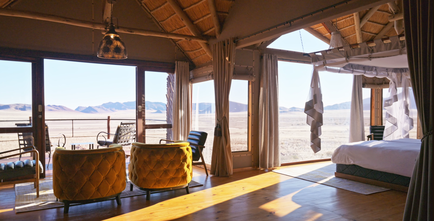 Luxurious safari lodge room with golden chairs, canopy bed, and panoramic desert views through open glass doors.
