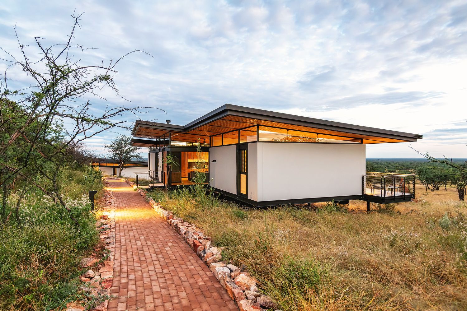 Contemporary eco-lodge with flat roof and large windows set in African savanna landscape at dusk with dramatic sky.