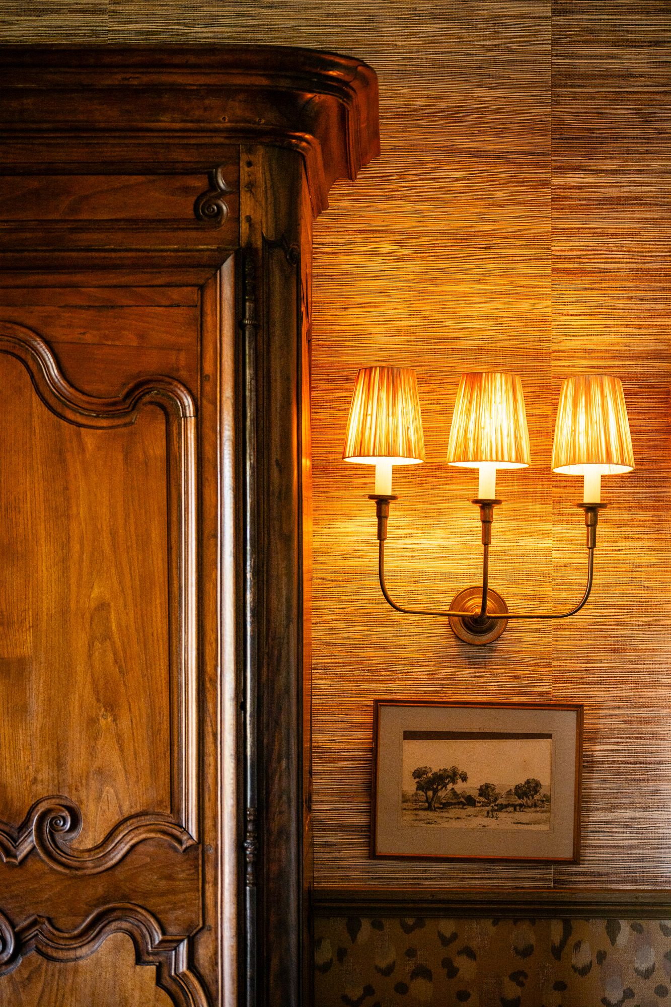 Elegant wall sconce with three glowing amber shades mounted on textured wallpaper beside ornate carved wooden armoire.