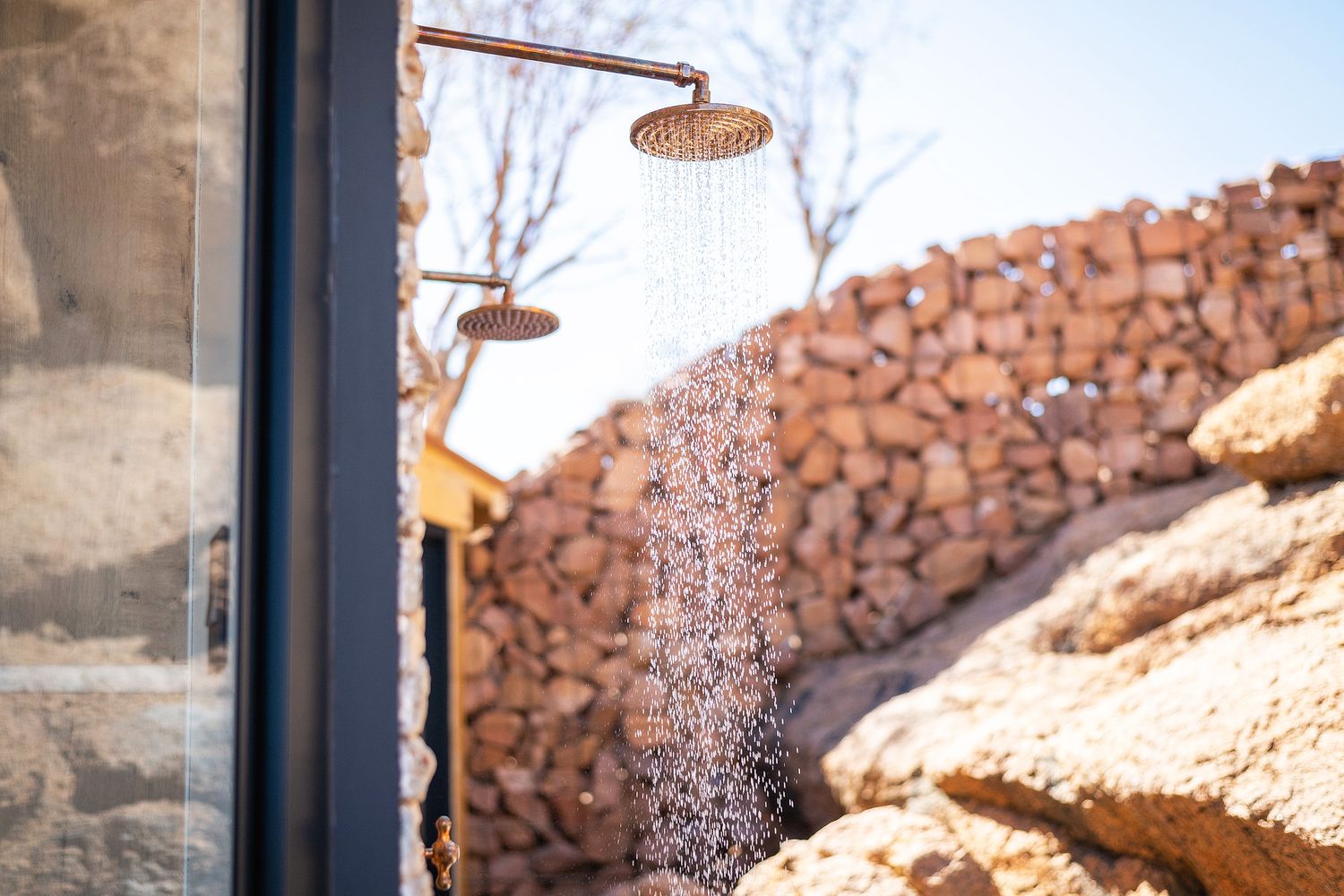 Outdoor shower with rain head mounted on stone wall, water flowing in natural rocky landscape setting.