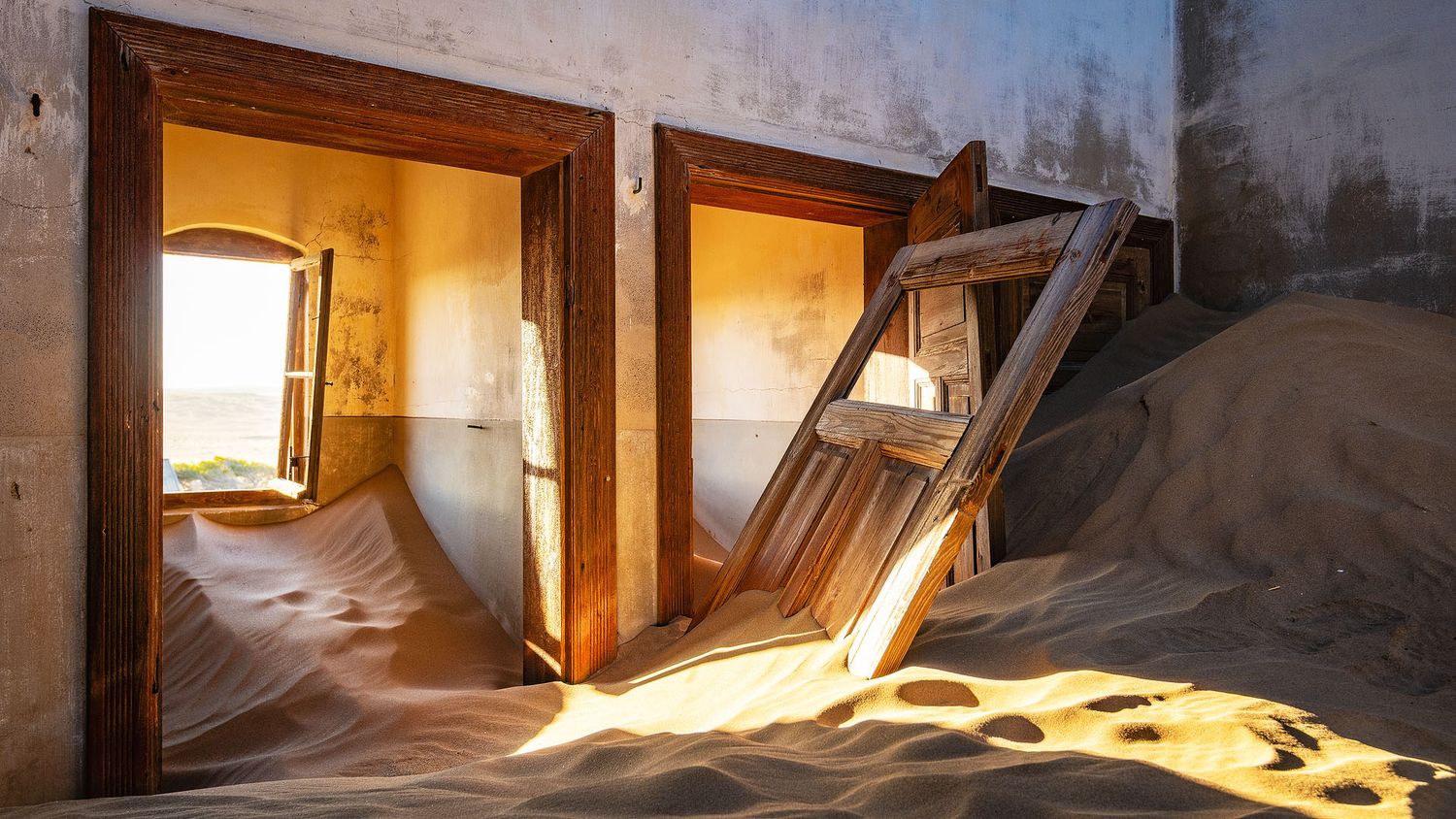 Abandoned desert building with sand-filled rooms, weathered wooden doors, and golden sunlight streaming through windows in Namibia.