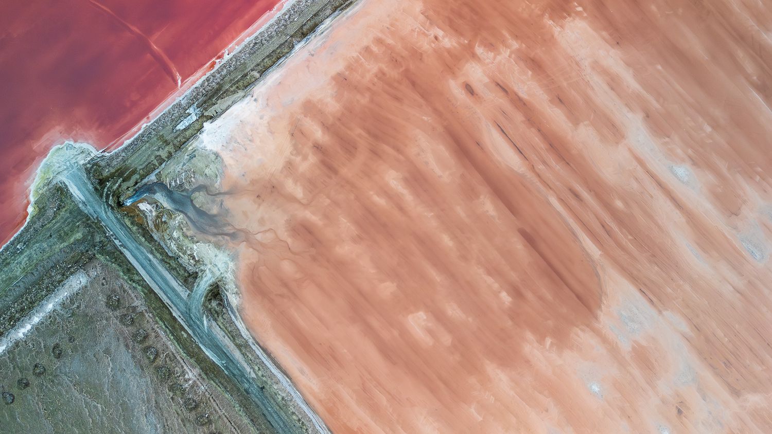 Aerial view of a pink salt lake meeting sandy shores, divided by a narrow road, showcasing vivid natural color contrasts.