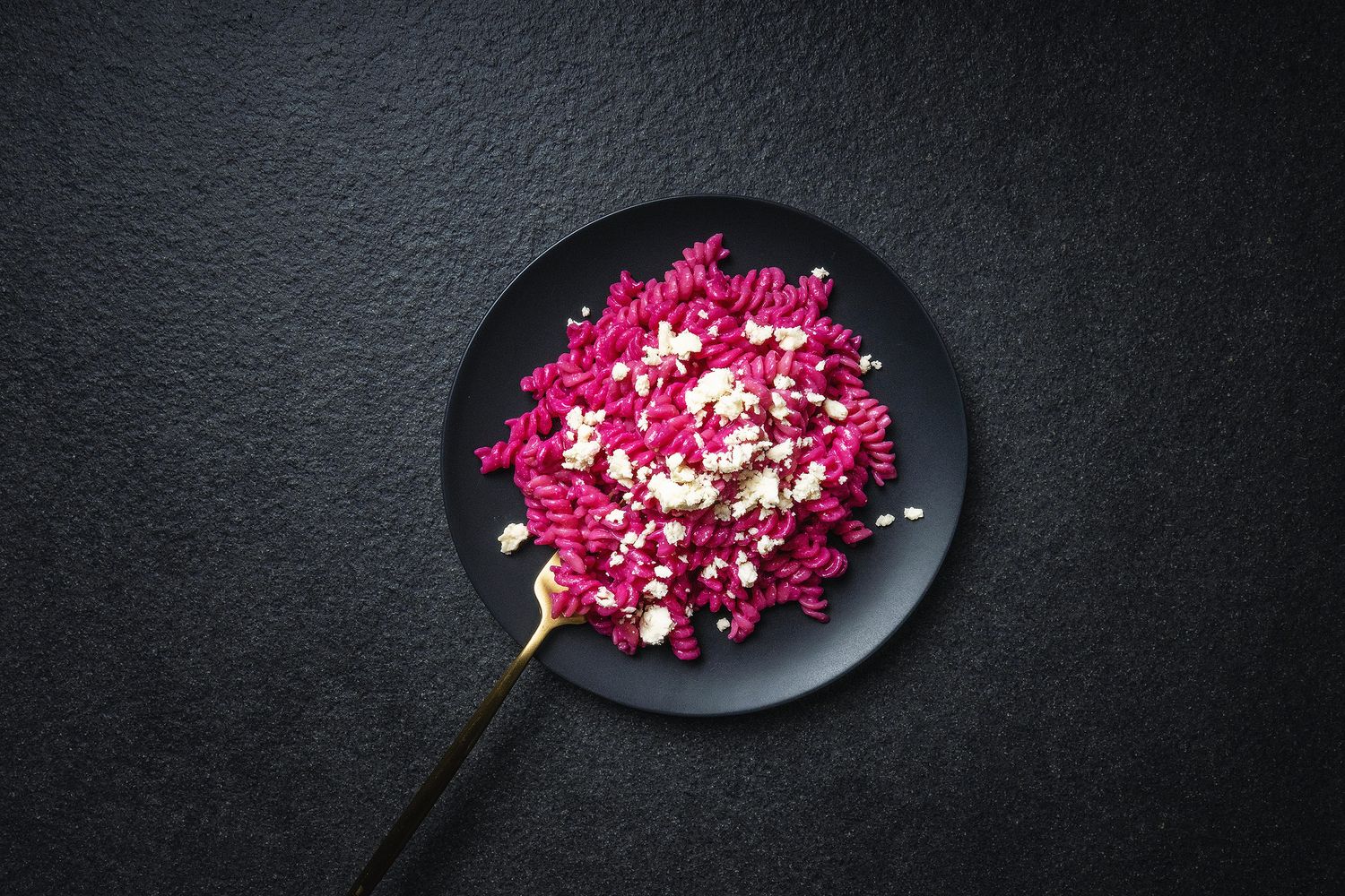 Pink fusilli pasta with crumbled white cheese served on a black plate with a gold fork on dark background.