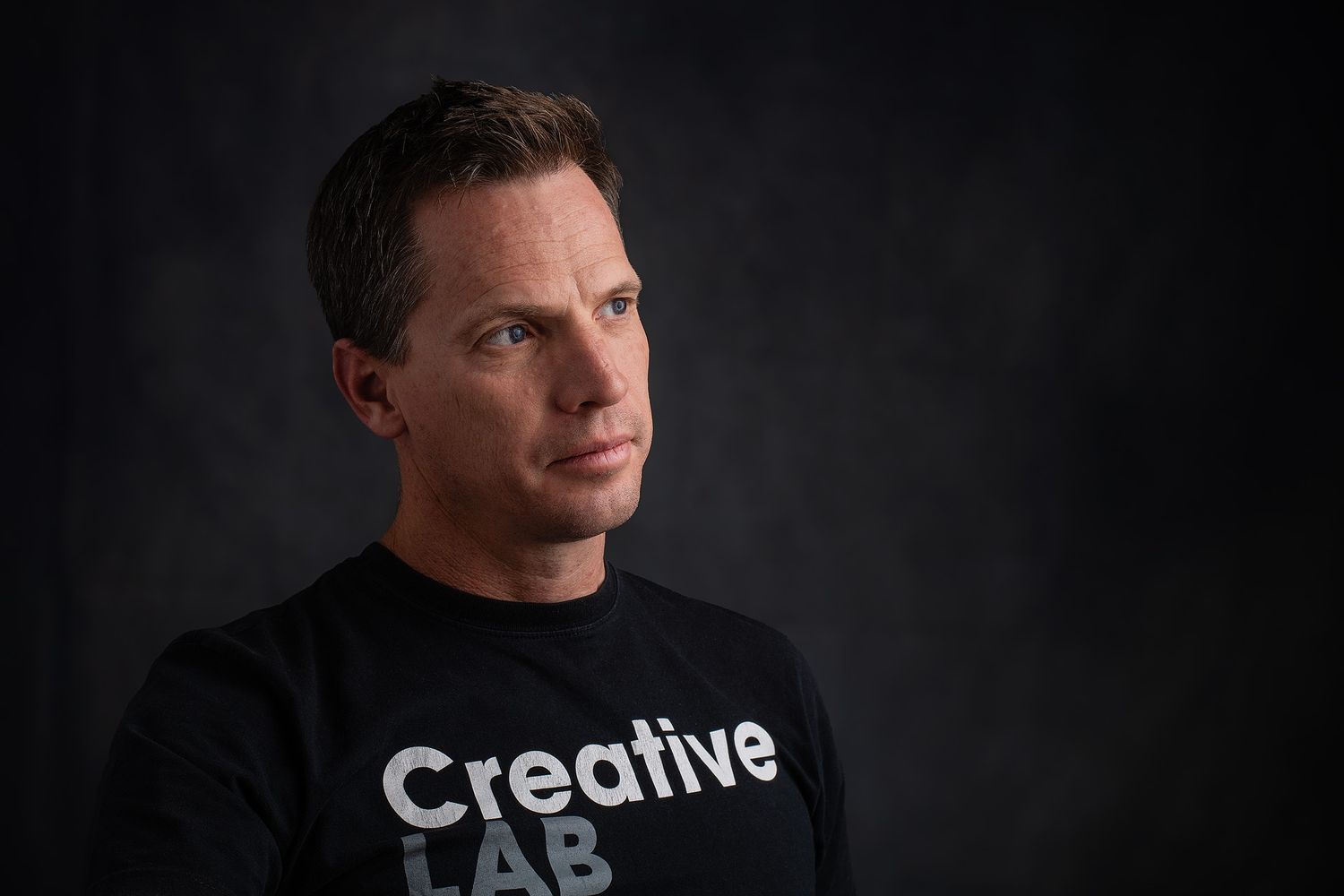 Man in black Creative Lab t-shirt poses against dark background in professional studio portrait photography.