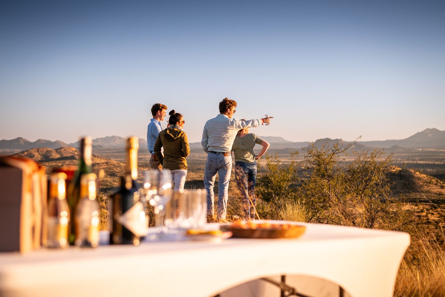 Group of tourists enjoying sundowner drinks at a scenic overlook with panoramic African landscape views.