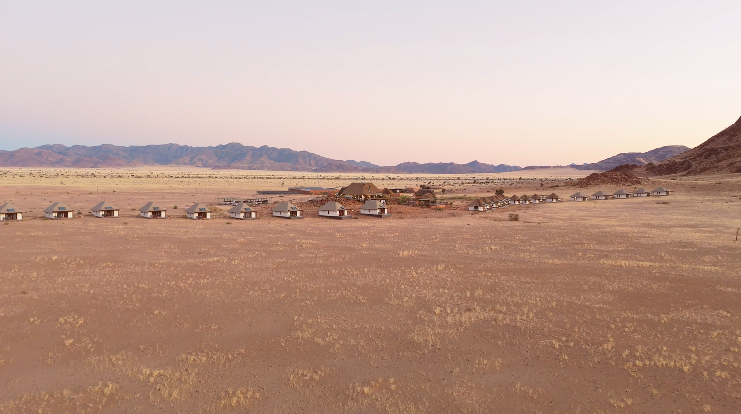 Aerial view of a remote desert lodge with scattered buildings against vast arid plains and mountain backdrop at dusk.