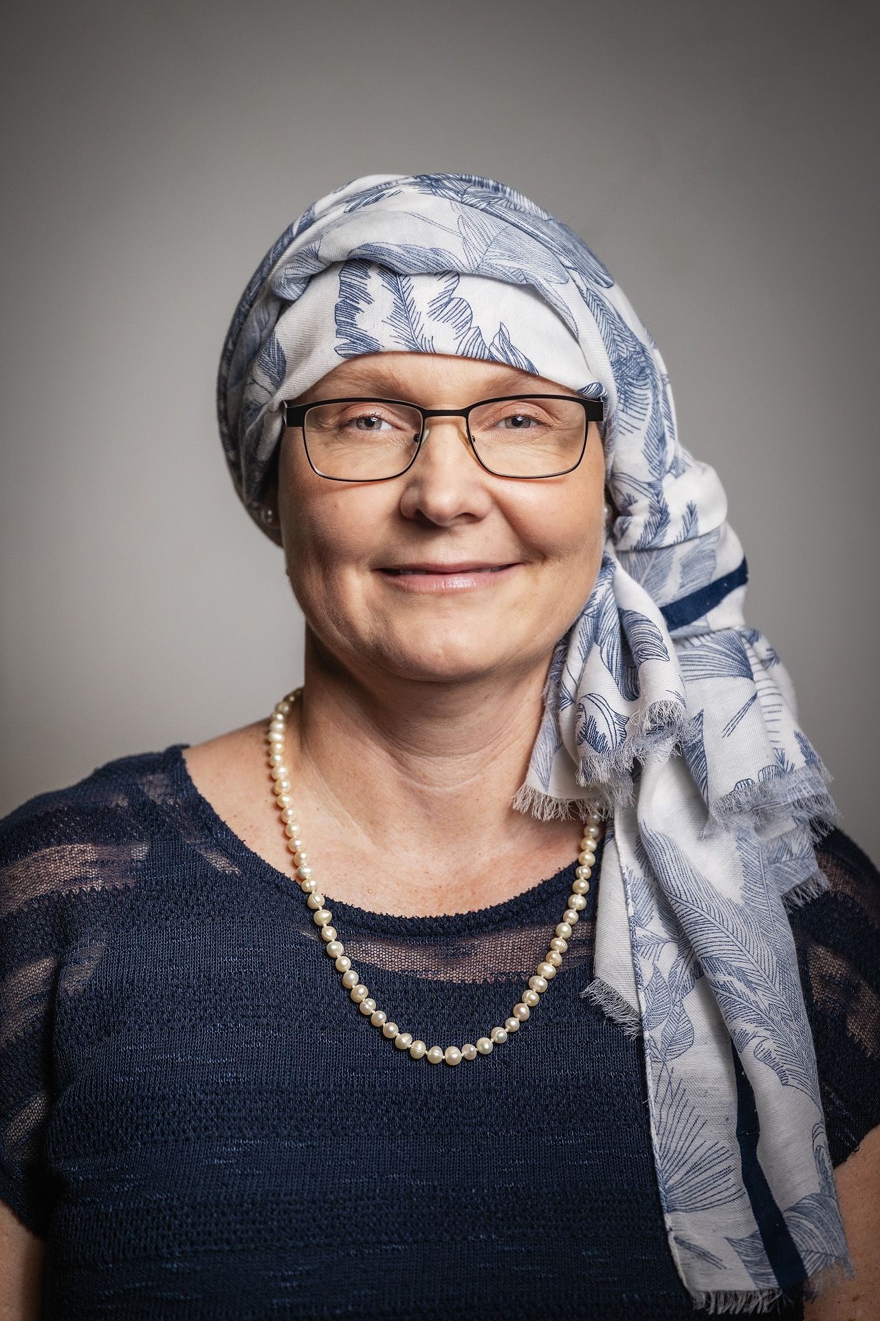 Smiling middle-aged woman wearing glasses, blue floral head scarf, pearl necklace, and navy top against gray background.