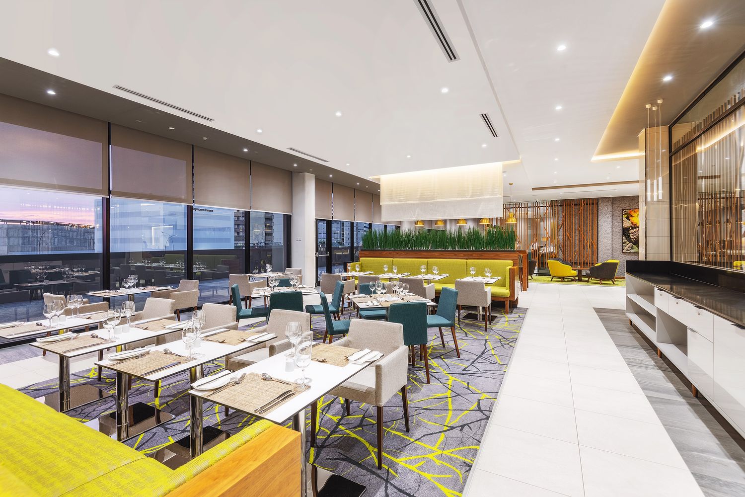 Modern hotel restaurant with elegant dining tables, teal chairs, yellow geometric carpet, and floor-to-ceiling windows with city views.