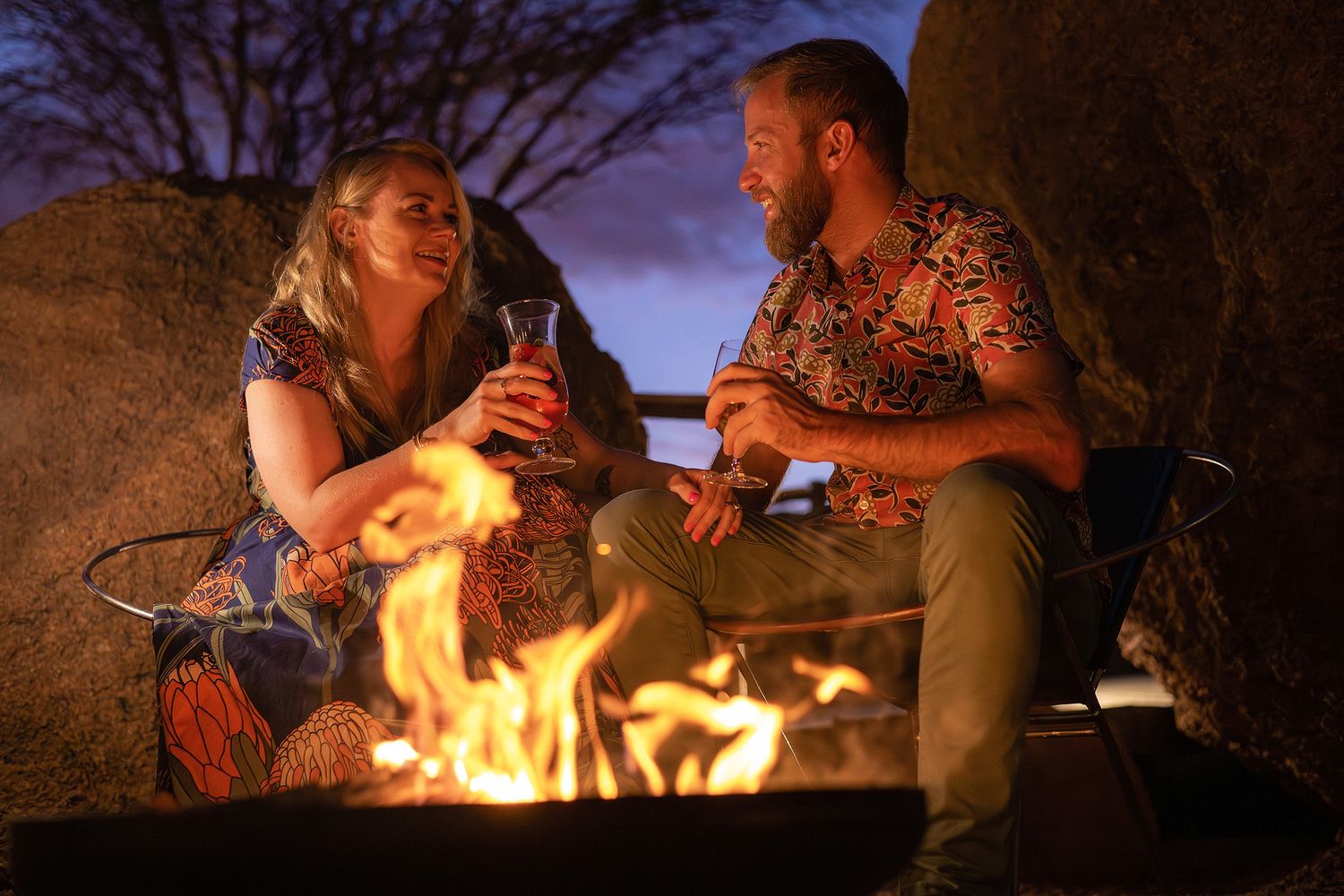 Couple toasting drinks by a campfire at dusk, surrounded by rocks in a warm outdoor setting.
