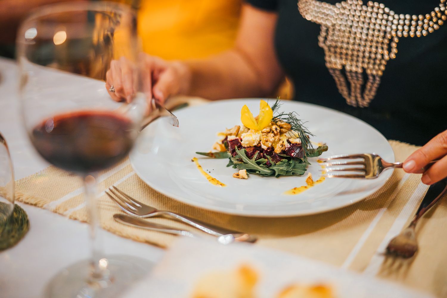 Elegant fine dining plate with gourmet salad and yellow garnish beside a glass of red wine on a set table.