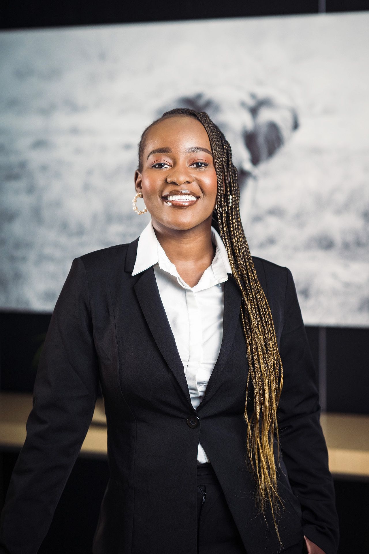 Smiling young Black businesswoman in black blazer and white shirt with braided hair stands before elephant artwork.