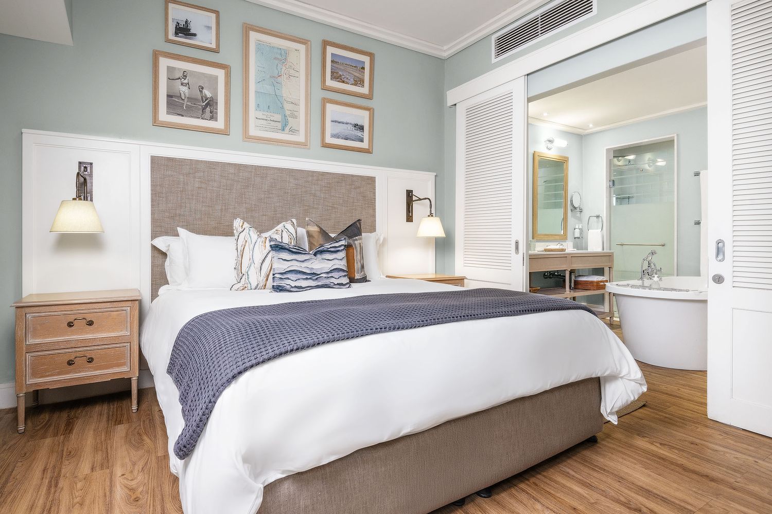 Elegant hotel bedroom with king bed, white bedding, navy throw, wood nightstands, and en-suite bathroom visible through open door.