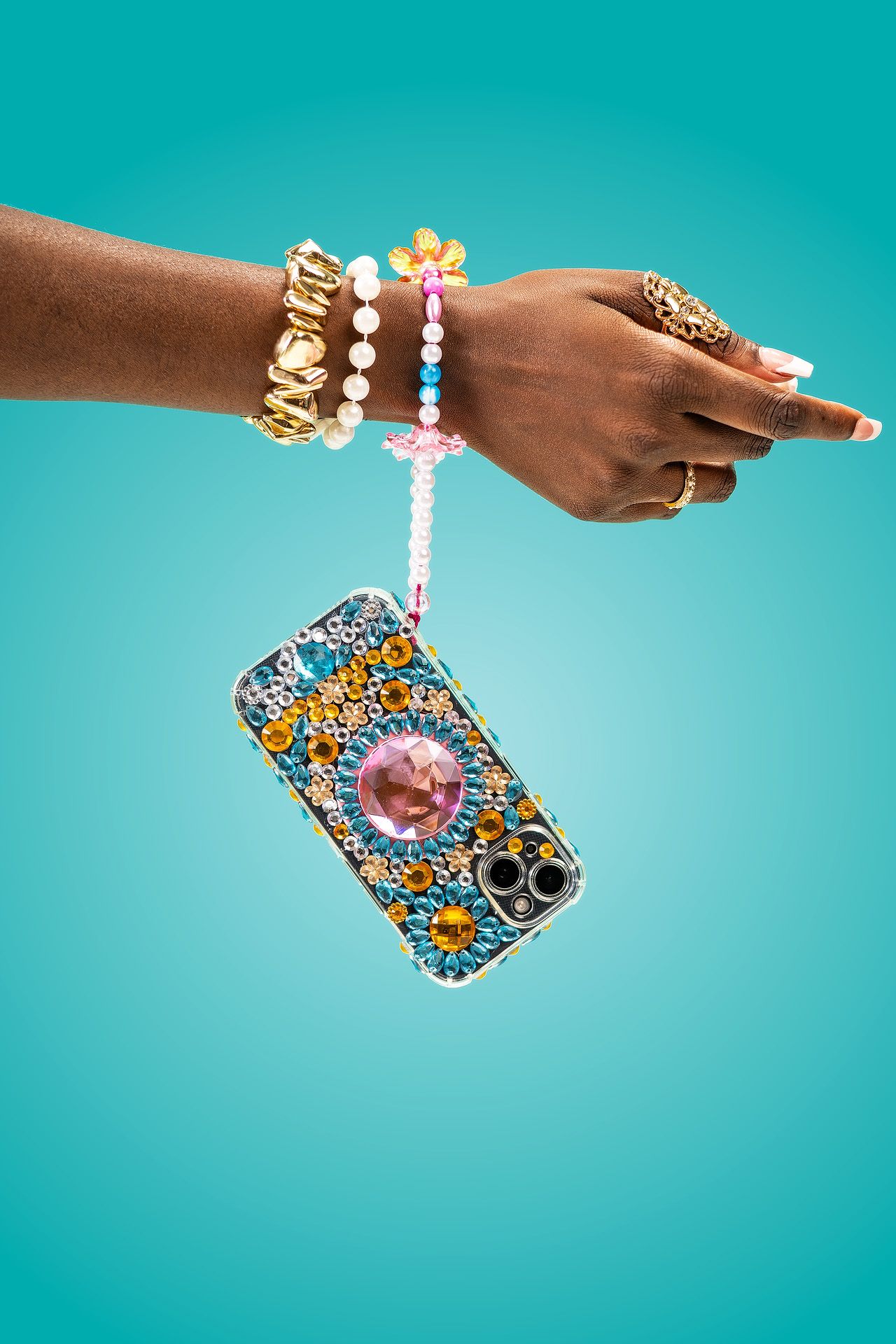Bejeweled floral phone case hanging from a beaded strap on a decorated hand with gold rings and bracelets.